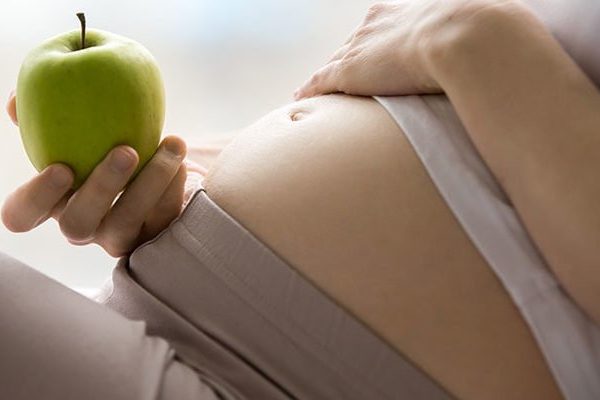 Eine schwangere Person hält einen grünen Apfel und berührt sanft ihren Bauch.