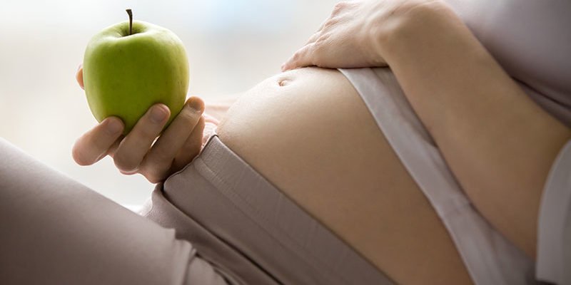 Eine schwangere Person hält einen grünen Apfel und berührt sanft ihren Bauch.
