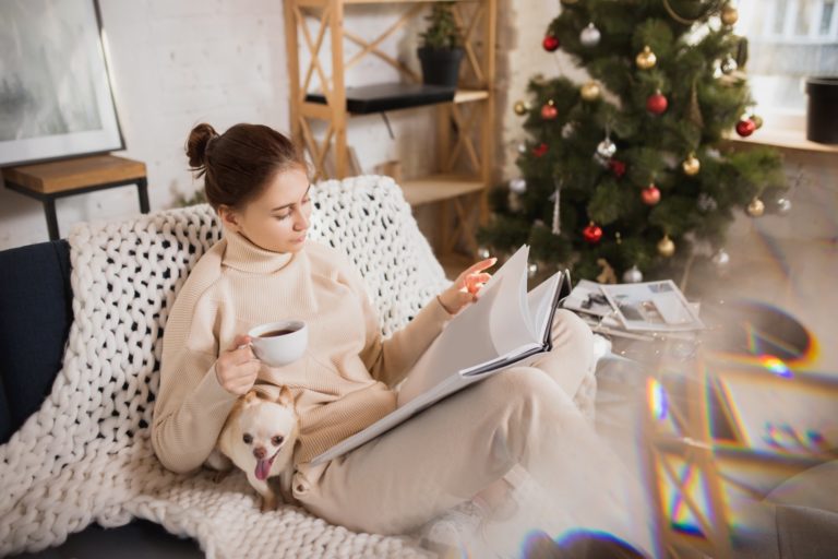 Eine Frau in bequemer Kleidung sitzt auf einem Sofa, liest in einem Buch und hält eine Tasse Kaffee, während ein Hund bei ihr sitzt.