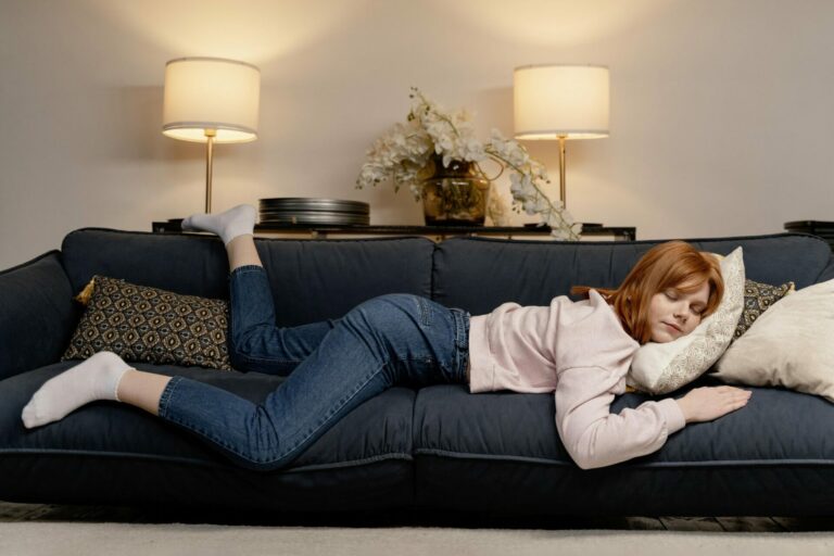 Eine junge Frau mit roten Haaren liegt entspannt auf einem blauen Sofa, umgeben von Kissen und Lampen.