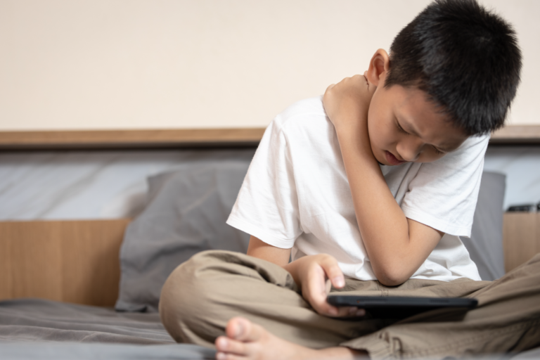 Ein Junge sitzt auf einem Bett, hält ein Tablet und fasst sich schmerzverzerrt an den Hals.